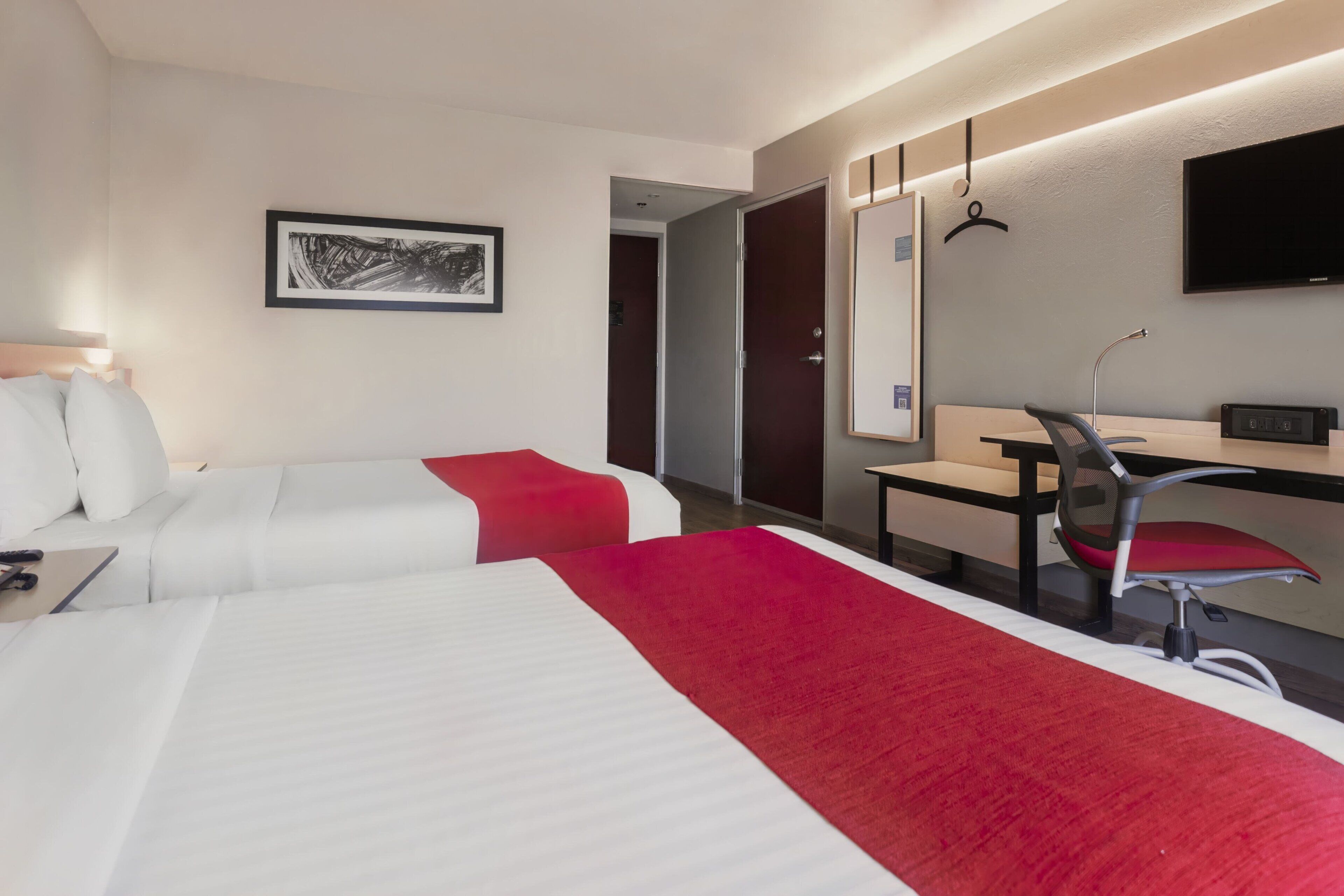 Habitación de hotel con dos camas, un escritorio, una silla, un televisor y una puerta que da a otra habitación.