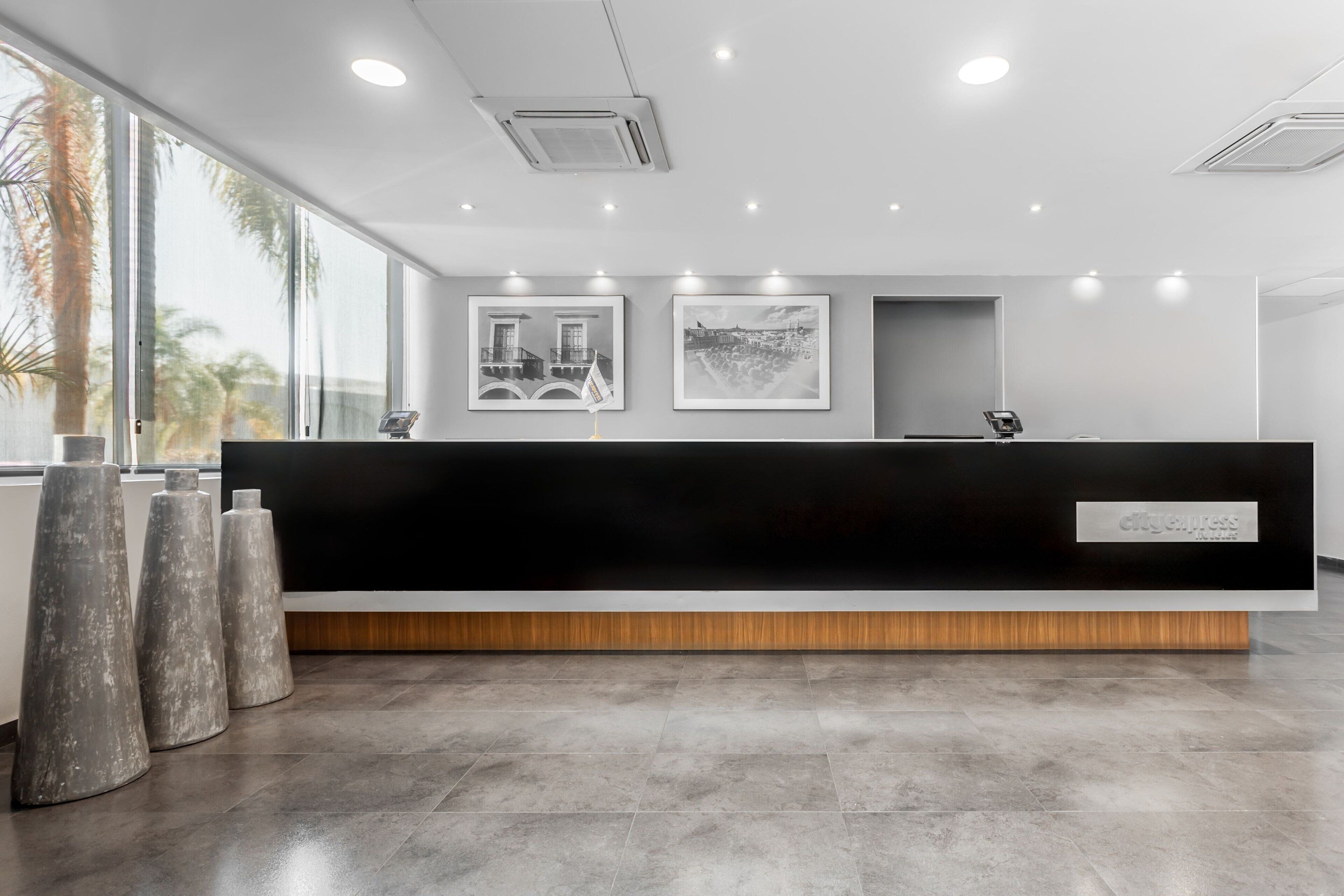 Un mostrador de recepción moderno con superficie negra y detalles de madera, flanqueado por dos taburetes grandes de forma cónica.