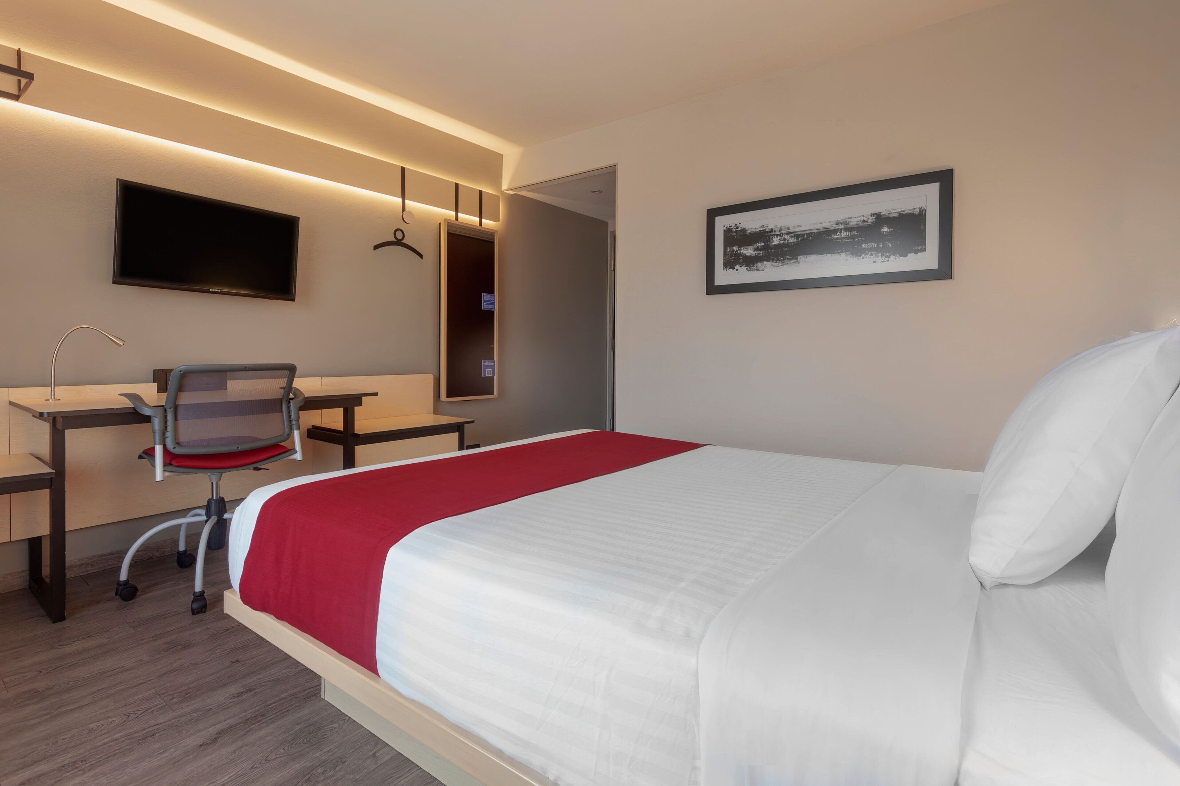 A hotel room with a bed, desk, chair, TV, and artwork.