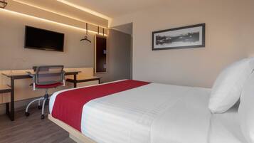 A hotel room with a bed, desk, chair, TV, and artwork.