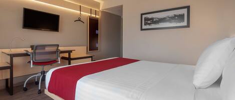 A hotel room with a bed, desk, chair, TV, and artwork.