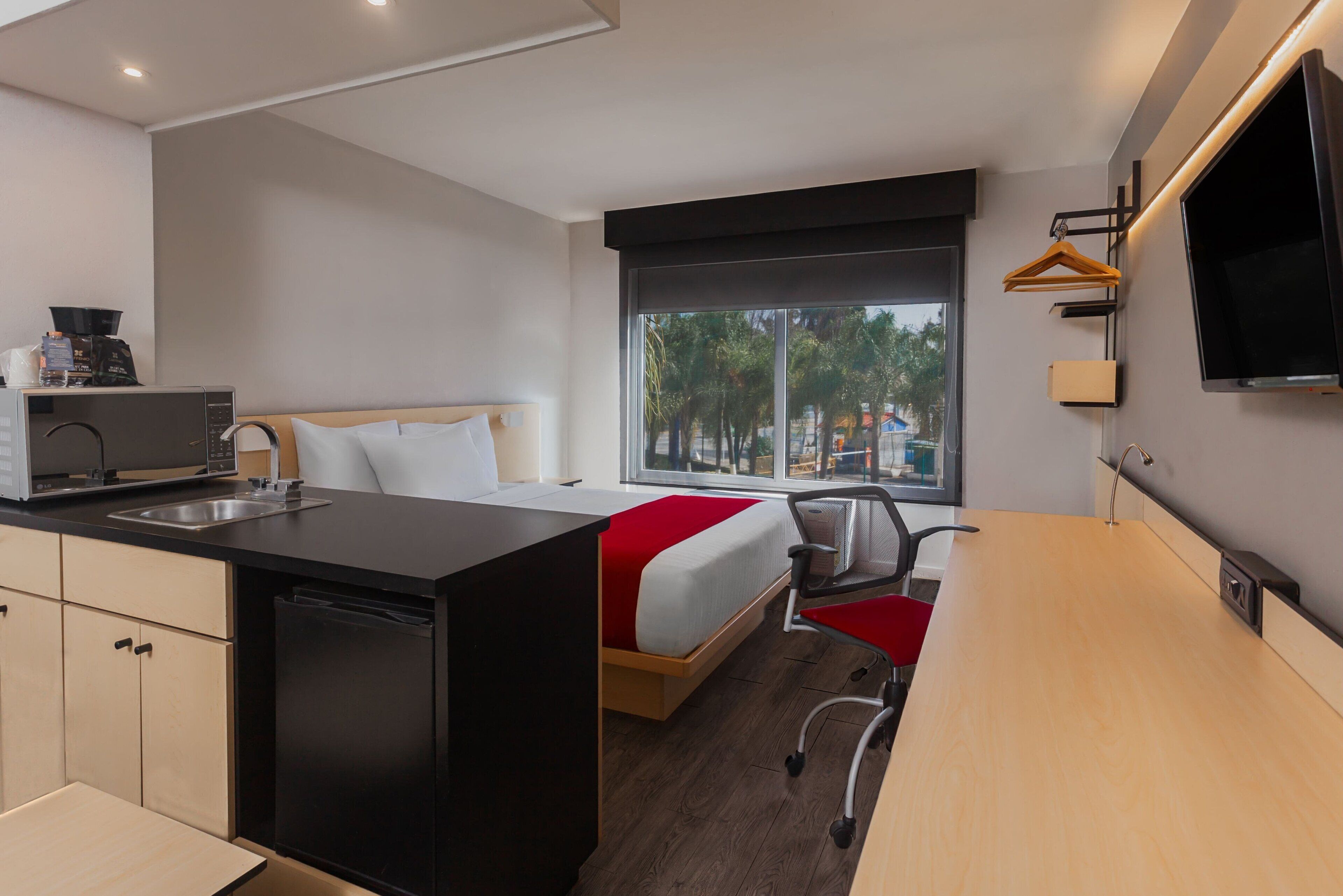 Habitación de hotel compacta con kitchenette, una cama con colcha roja y blanca, un escritorio y un amplio ventanal con vistas a palmeras y edificios.
