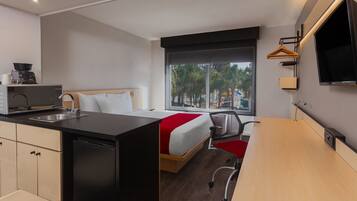 Habitación de hotel compacta con kitchenette, una cama con colcha roja y blanca, un escritorio y un amplio ventanal con vistas a palmeras y edificios.
