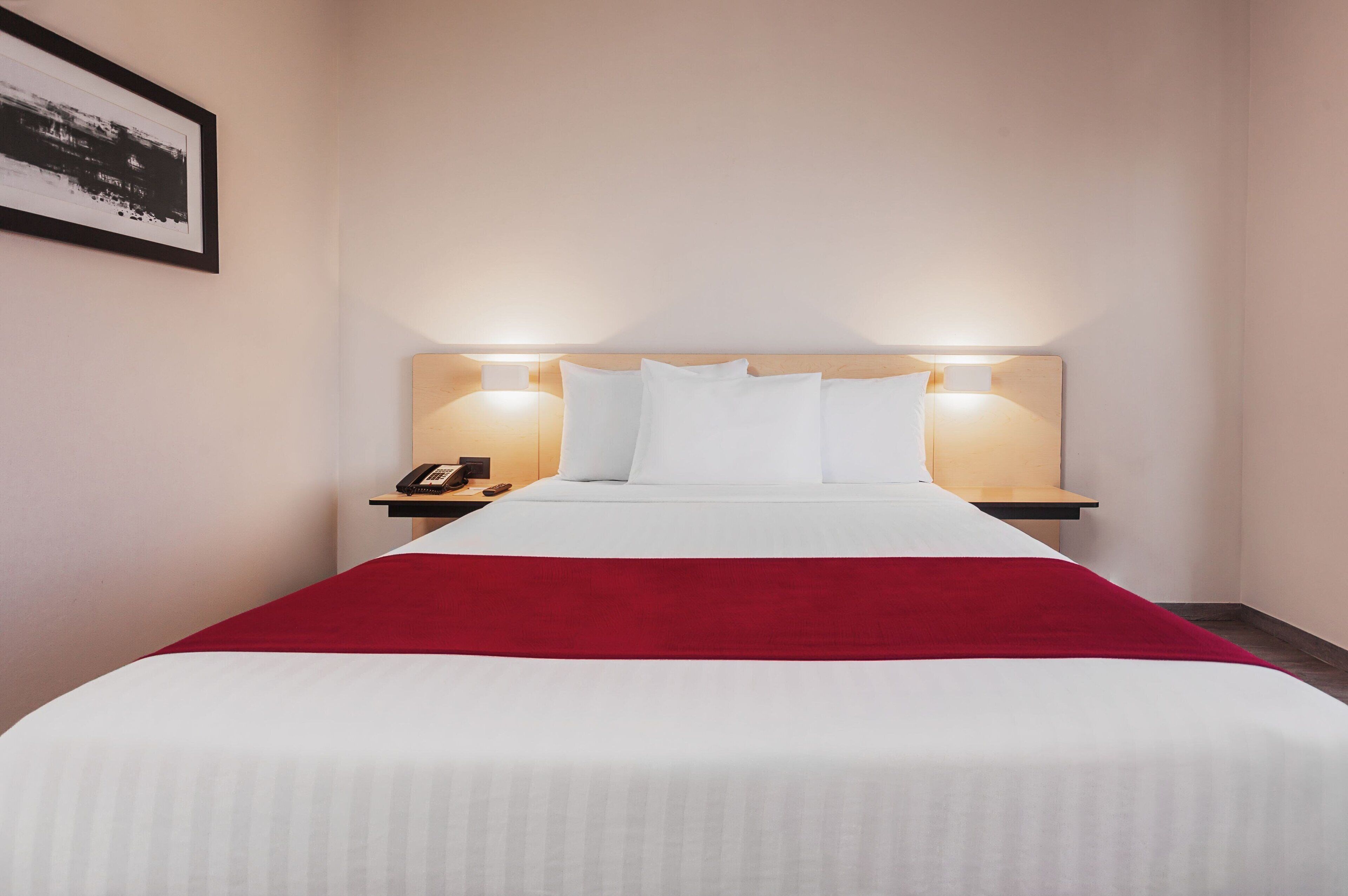 A hotel room with a neatly made bed, a red throw blanket, and bedside lamps.