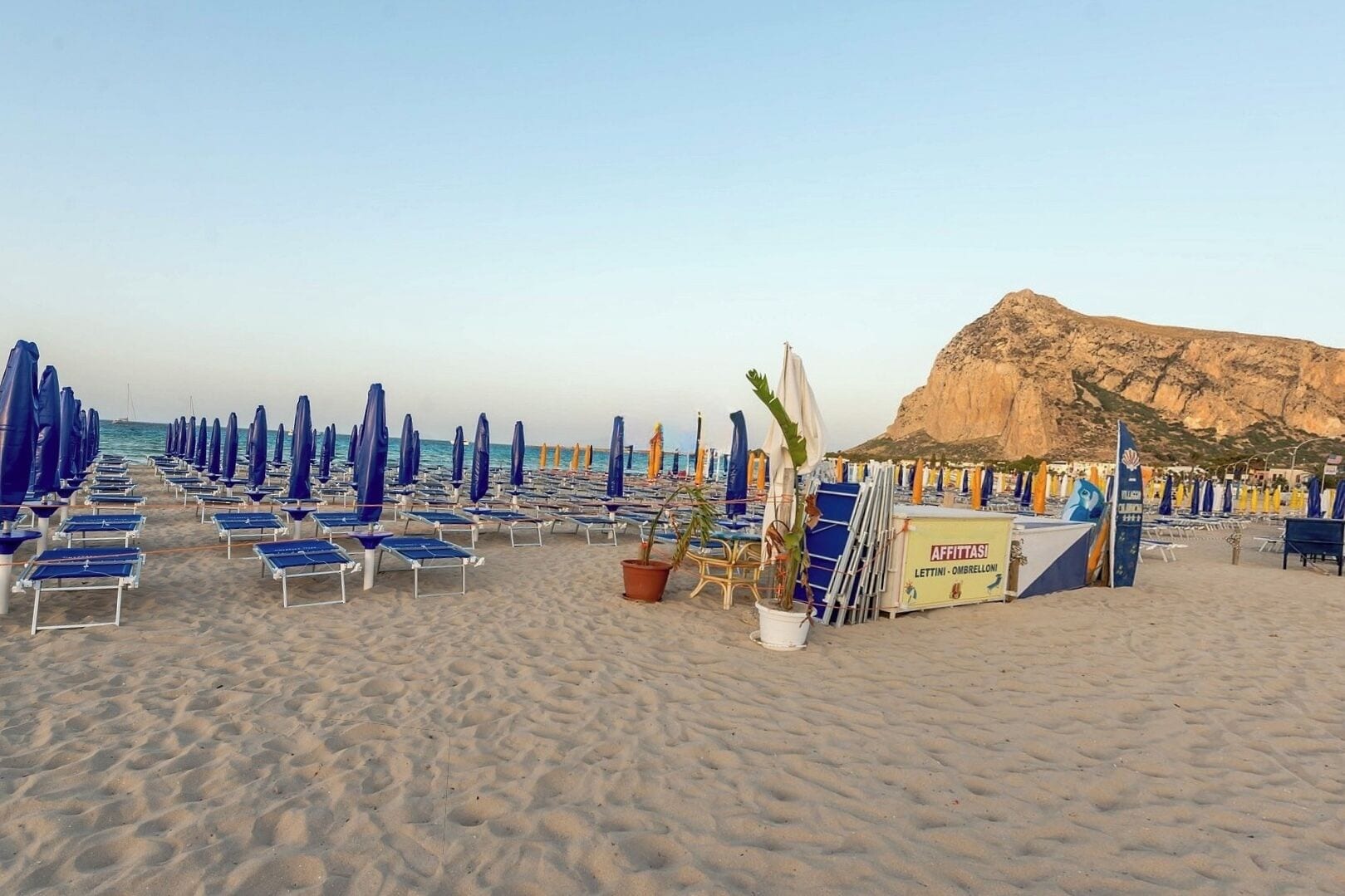A beach with rows of blue and white sun loungers and umbrellas, a rocky hill in the background, and a sign for 'AFFITTASI LETTINI - OMBRELLONI'.