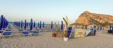 A beach with rows of blue and white sun loungers and umbrellas, a rocky hill in the background, and a sign for 'AFFITTASI LETTINI - OMBRELLONI'.