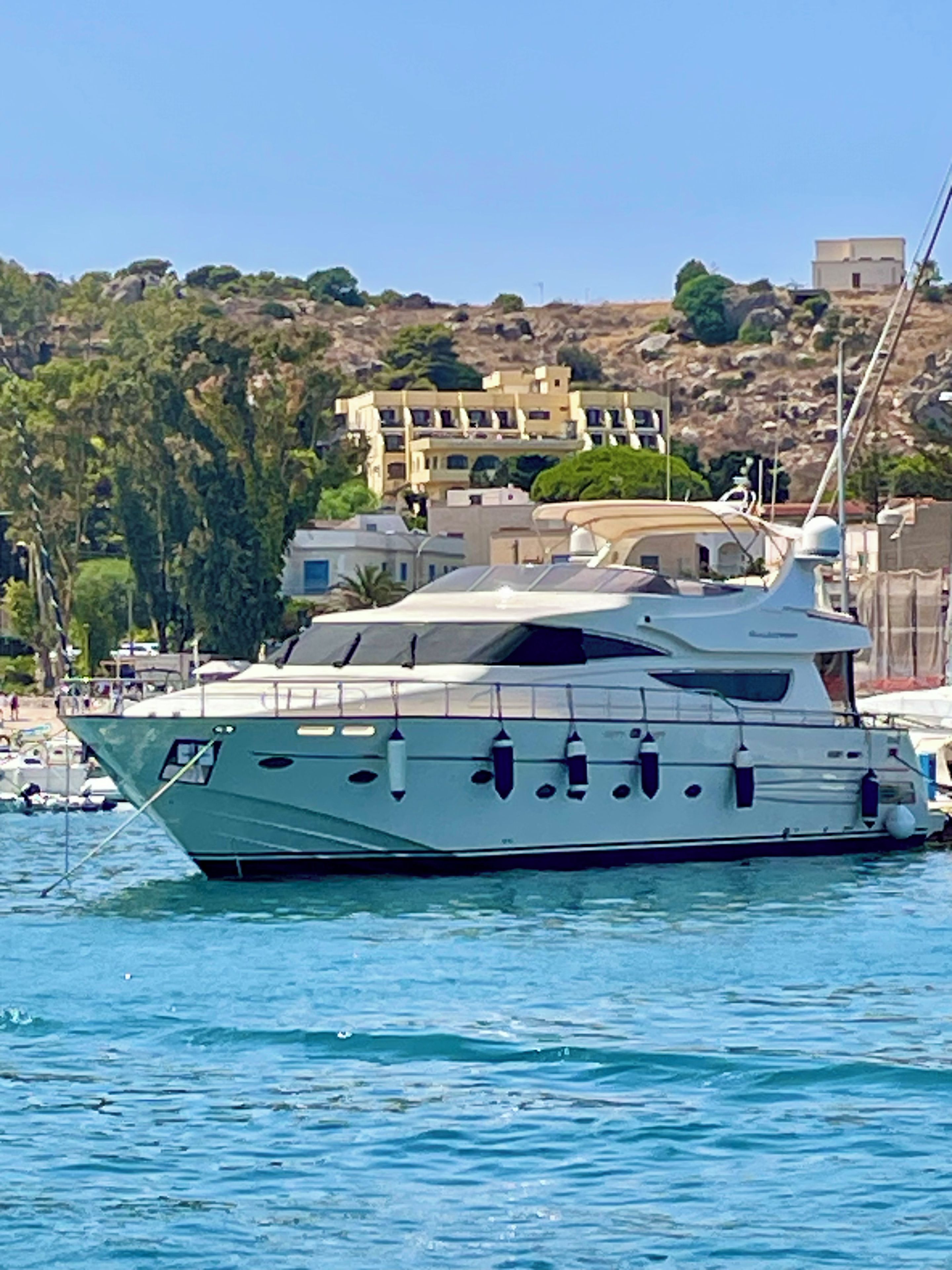 Un yate grande amarrado en un puerto deportivo con edificios y colinas al fondo.