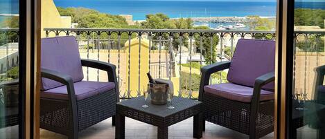 A balcony with two chairs and a table, overlooking a coastal view with buildings and the sea.