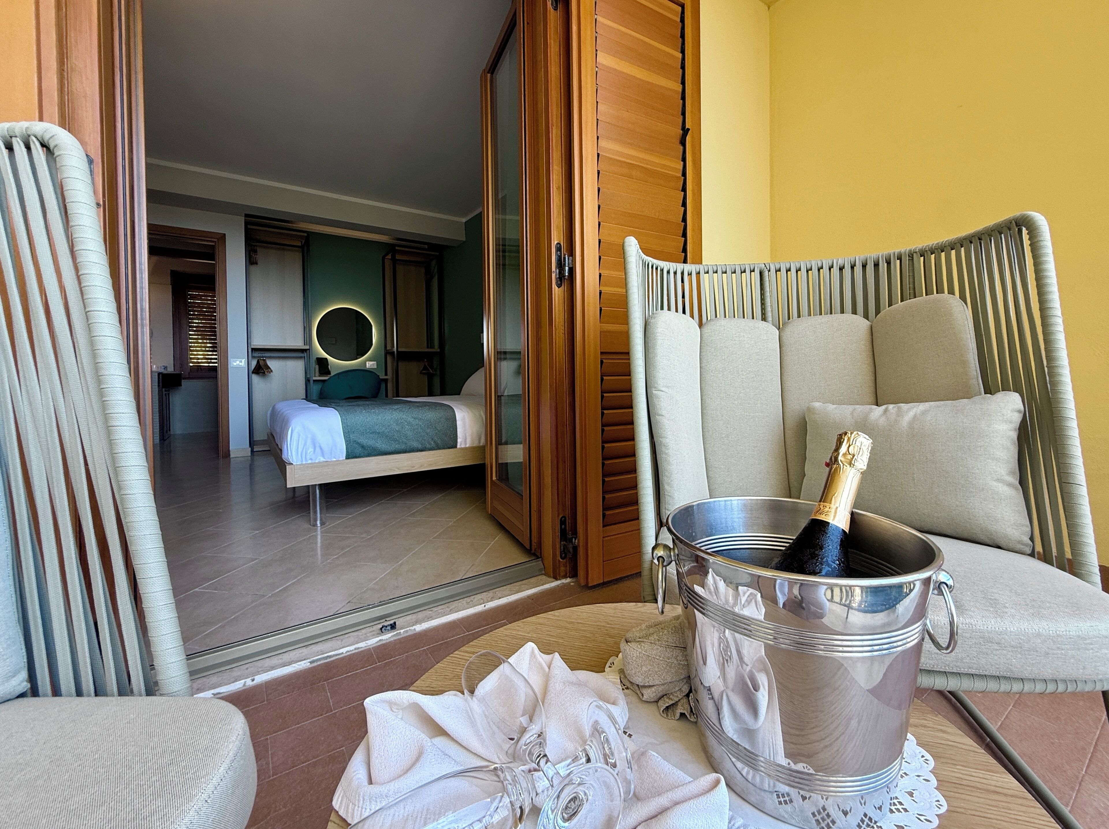 Una camera d'albergo con un letto, una poltrona, un divano, un minibar e una bottiglia di champagne.