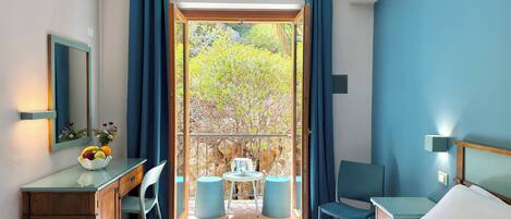 A hotel room with a bed, a desk with a mirror, a small table with chairs, and a balcony with a view of trees.