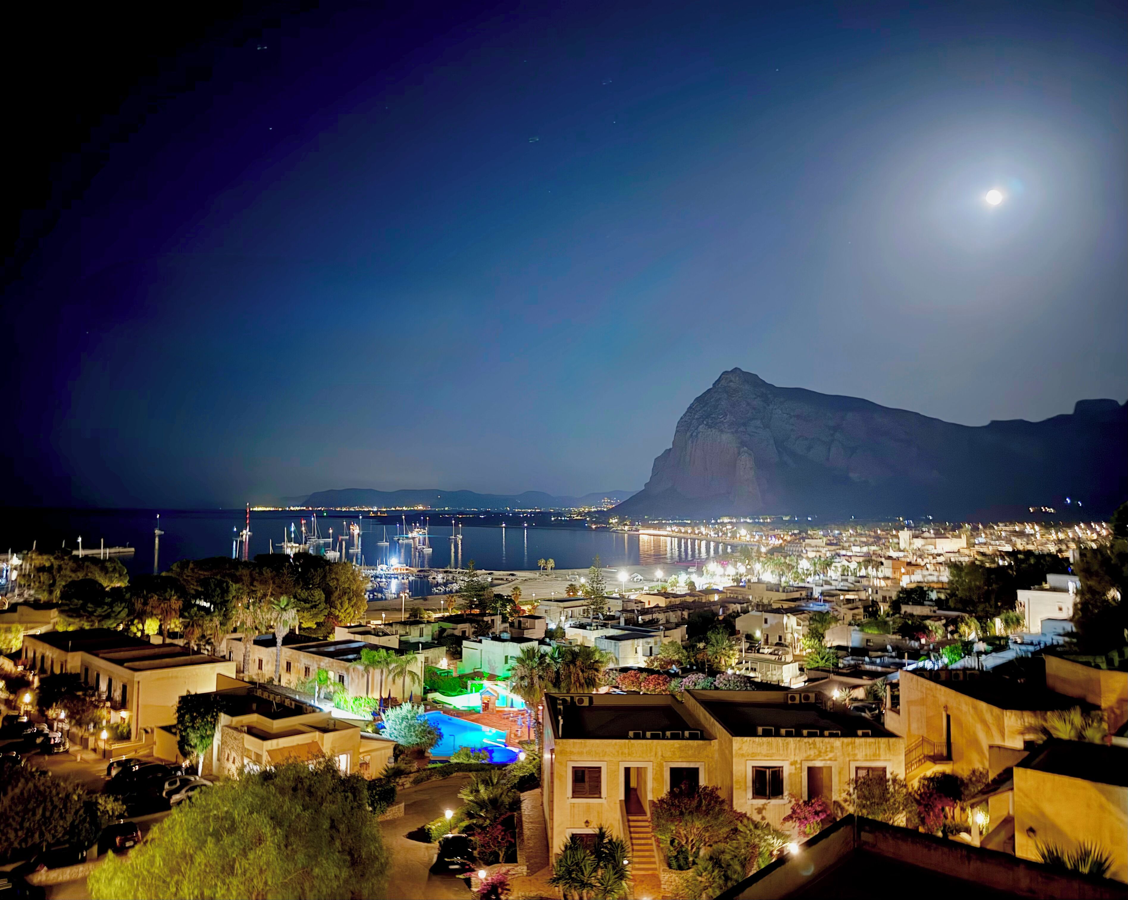 A coastal town at night with illuminated buildings, a marina, and a prominent mountain in the background.