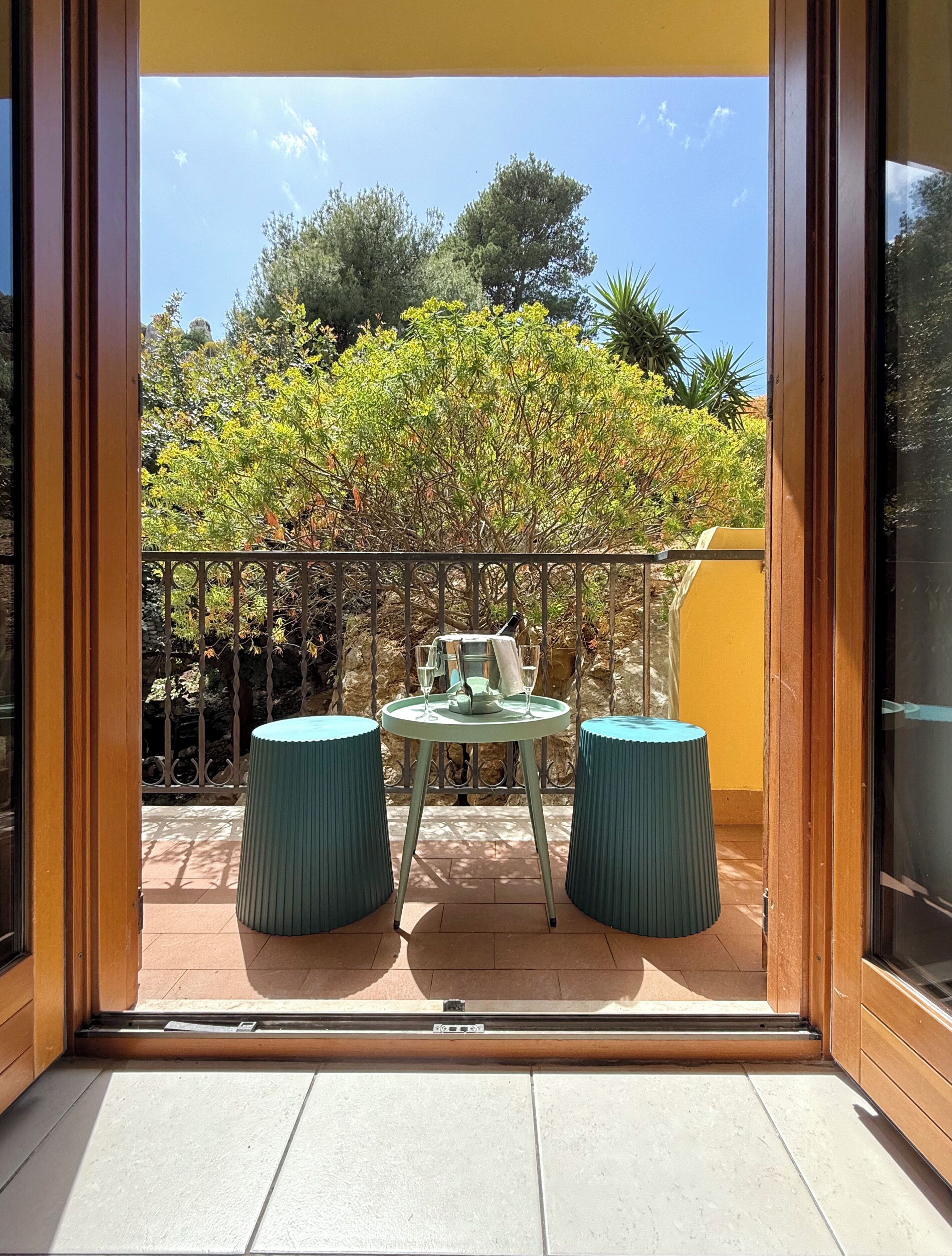 Un balcon avec une table et deux tabourets, une rambarde en métal et une vue sur les arbres et un ciel dégagé.