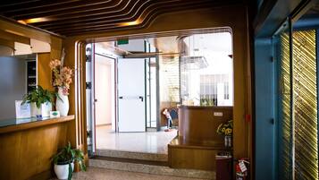 Un área de recepción con un mostrador de madera, plantas en macetas, un extintor y vistas al exterior a través de una puerta de cristal.