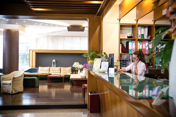 A reception area with a receptionist, a reception desk, a seating area with a sofa and chairs, and a bookshelf with binders. - Wyndham Garden Conegliano (Conegliano)
