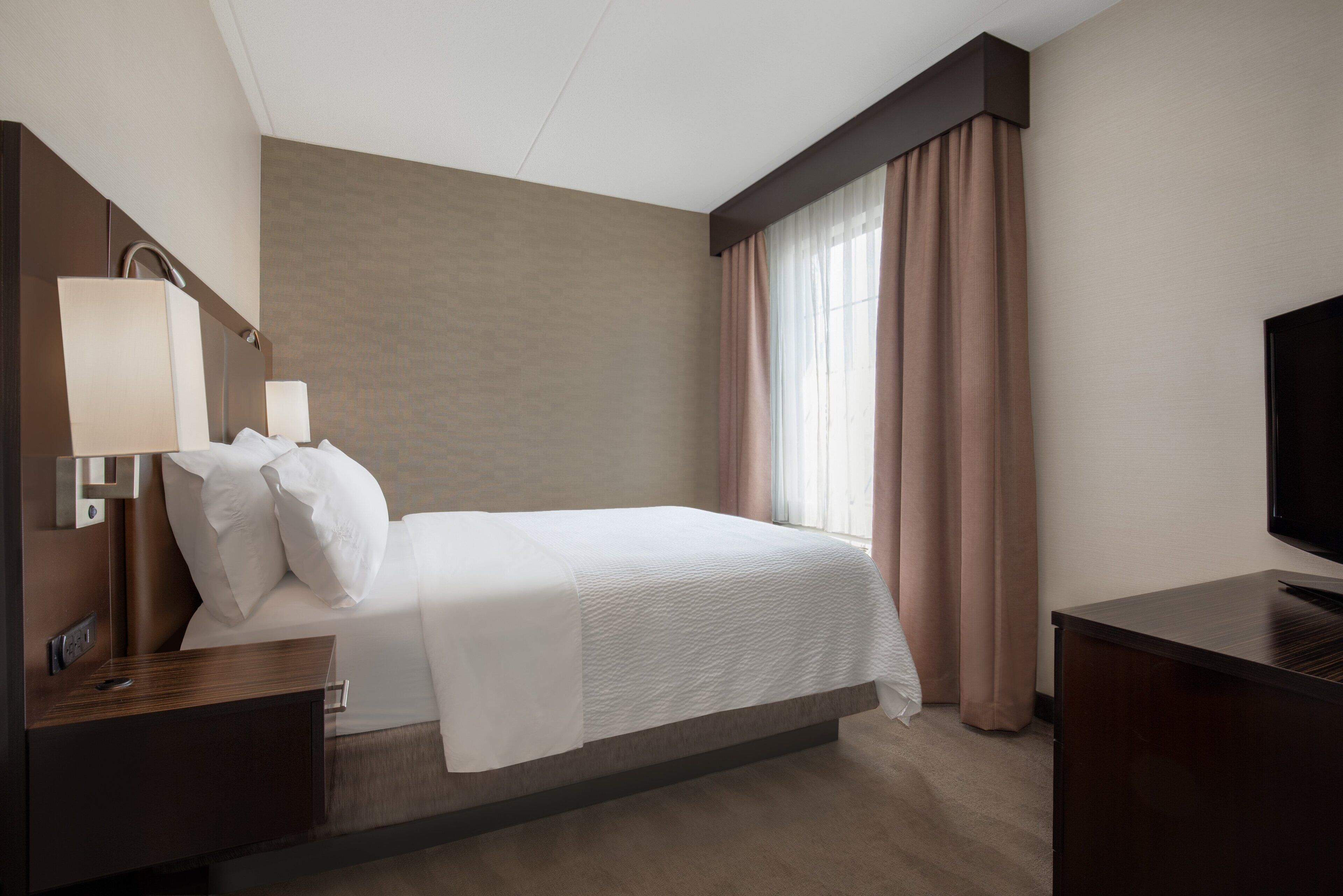 Una habitación de hotel con cama, mesita de noche, televisión y ventana con cortinas.