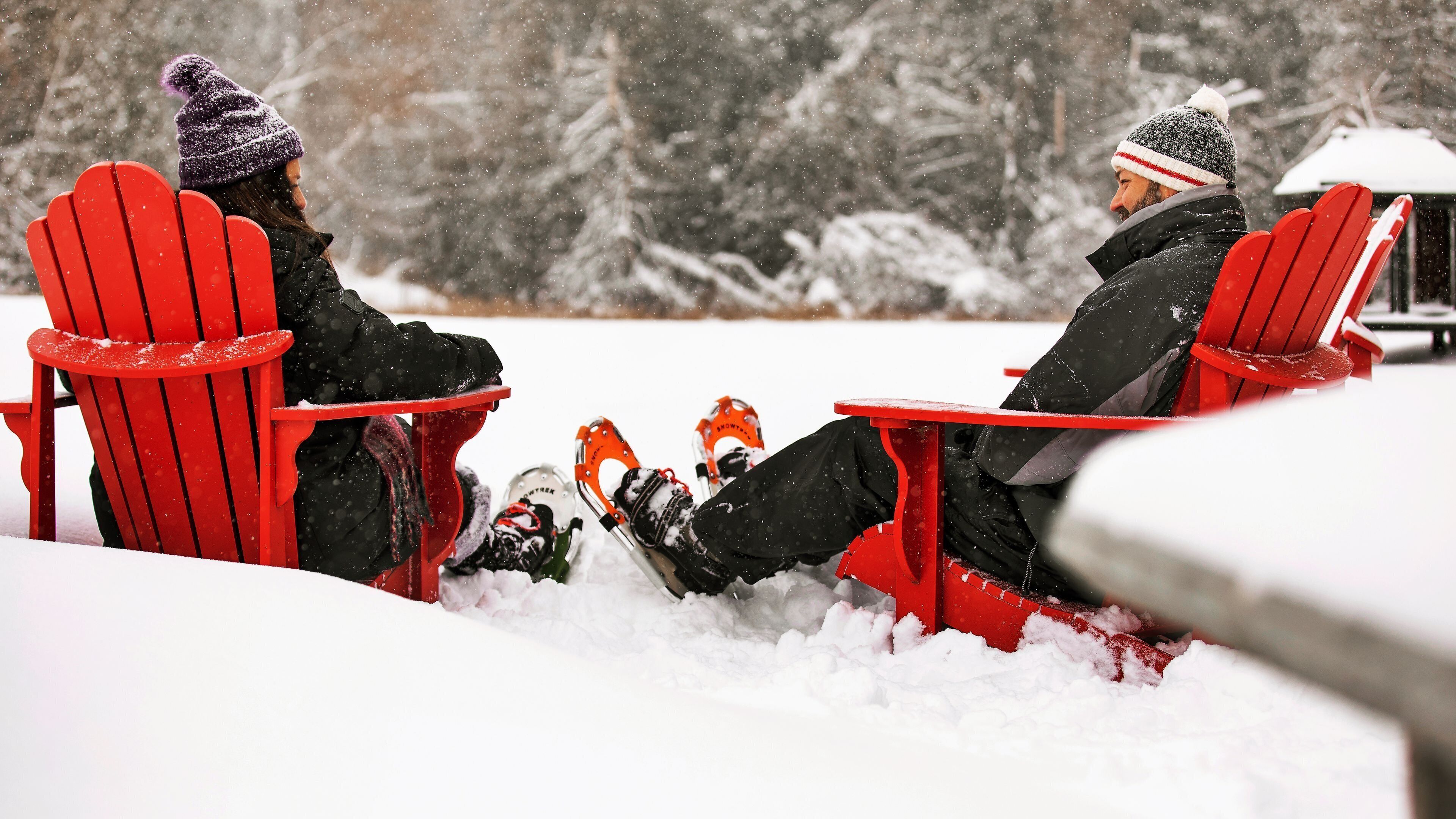 Two people seated on red Adirondack chairs in a snowy outdoor setting.