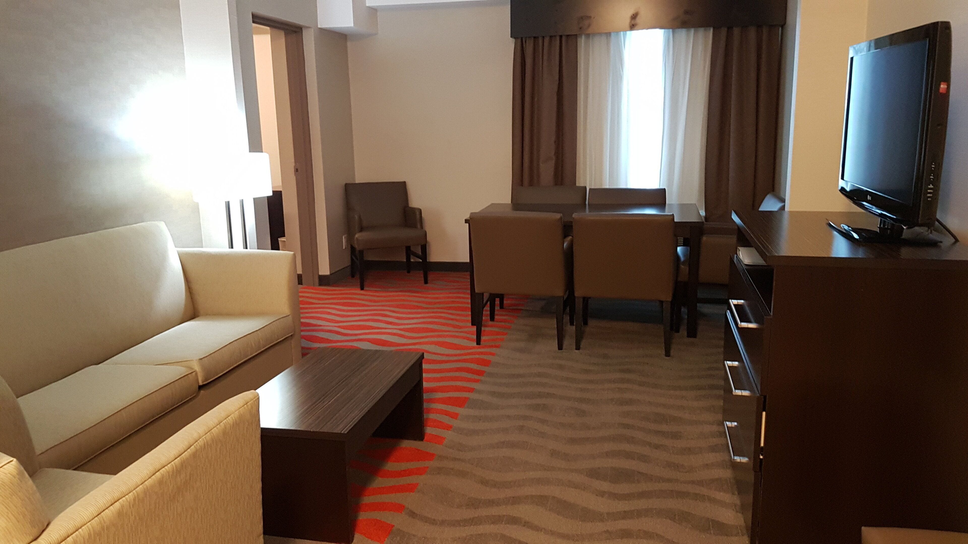 A hotel room with a sofa, a dining table with chairs, a television, and a patterned carpet.