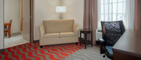 A hotel room with a sofa, a desk, a chair, a lamp, and a view of the outdoors through a window.
