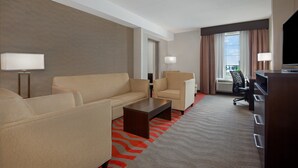 A hotel room with a beige sofa, two armchairs, a wooden coffee table, a desk, and a TV.