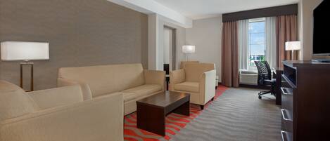 Habitación de hotel con sofá color beige, dos sillones, una mesa de centro de madera, un escritorio y un televisor.