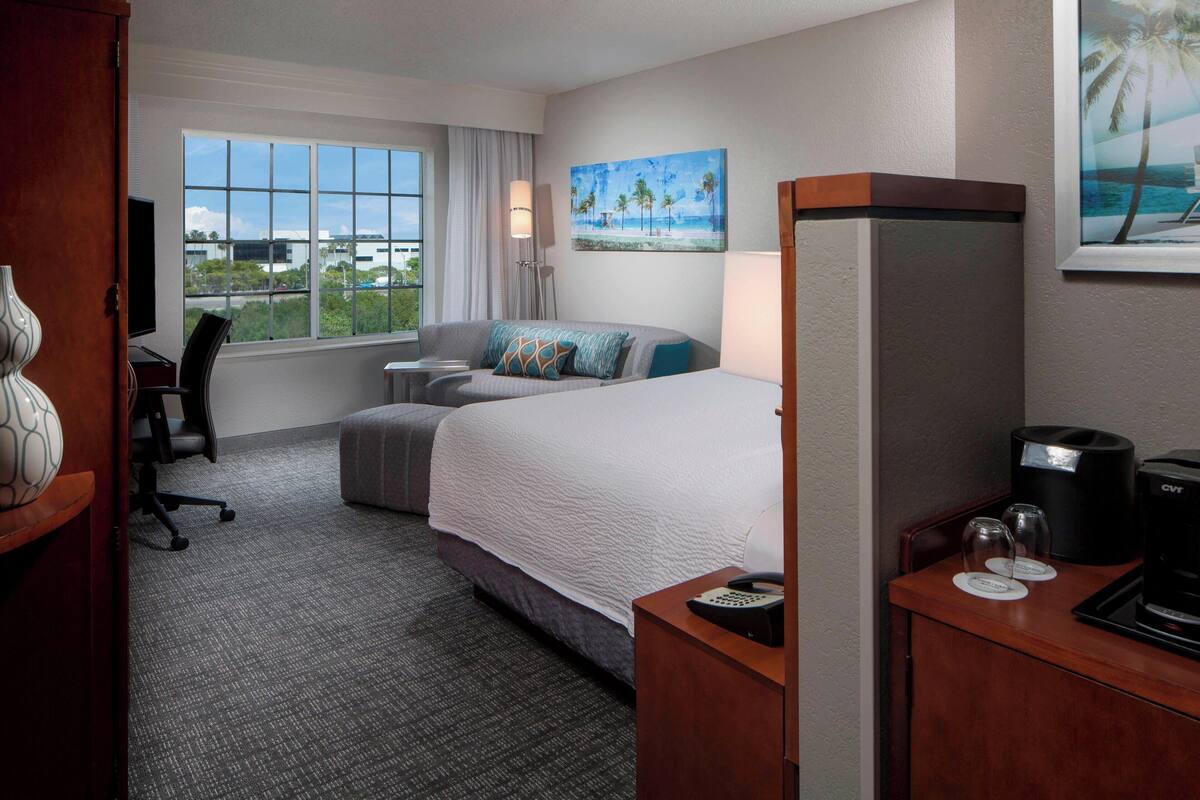A hotel room with a bed, a desk with a coffee maker, a chair, a TV, and a window with a view of palm trees and the ocean.