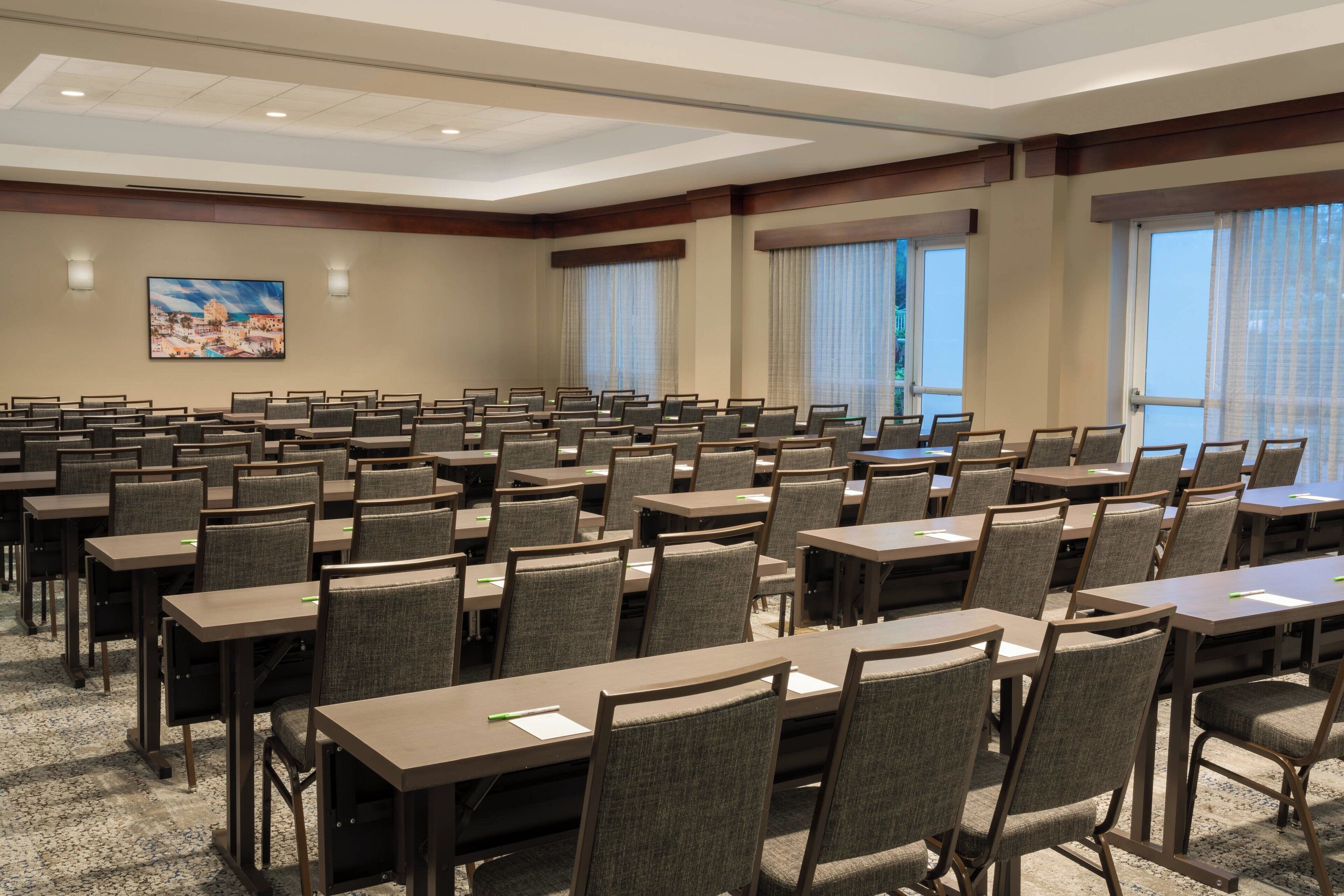 A conference room with rows of tables and chairs, large windows with curtains, and a painting on the wall.