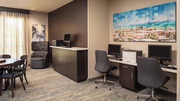A hotel room with a desk, two monitors, a printer, a chair, a dining table with chairs, a large window with curtains, and a cityscape painting on the wall.
