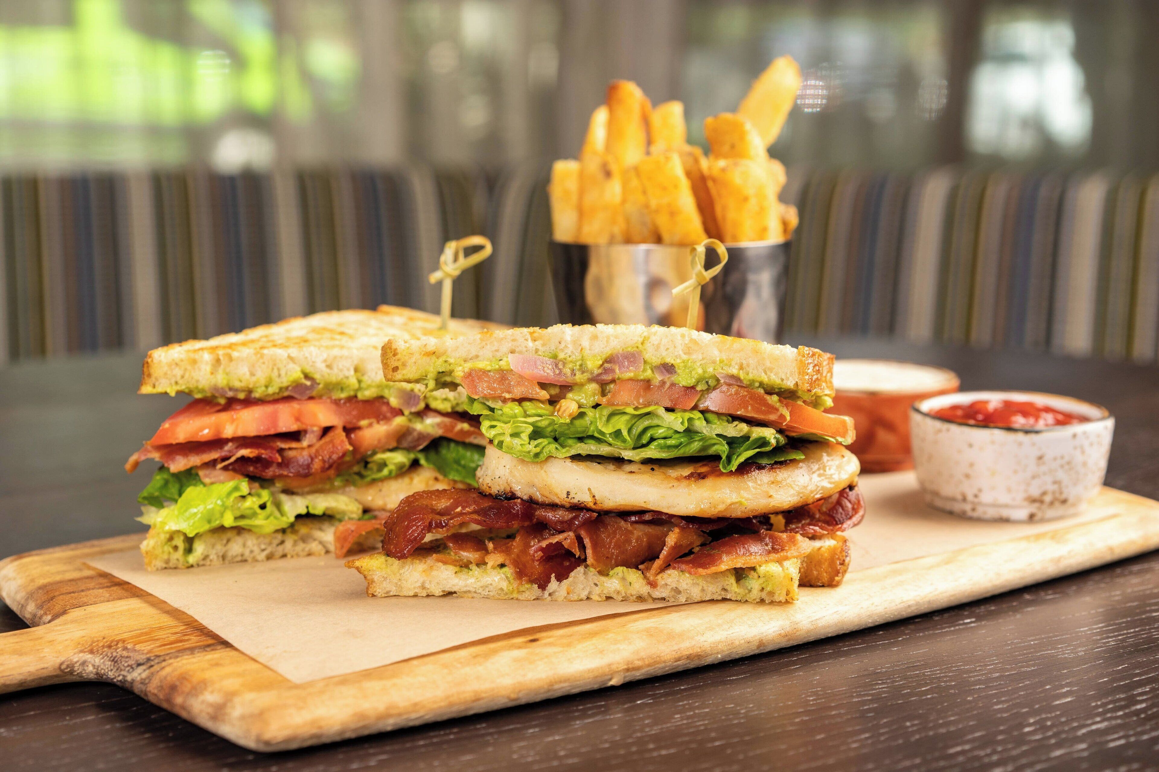 Sándwich de tocino, lechuga, tomate y queso sobre una tabla de madera, acompañado de papas fritas y kétchup.