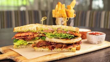 Sándwich de tocino, lechuga, tomate y queso sobre una tabla de madera, acompañado de papas fritas y kétchup.