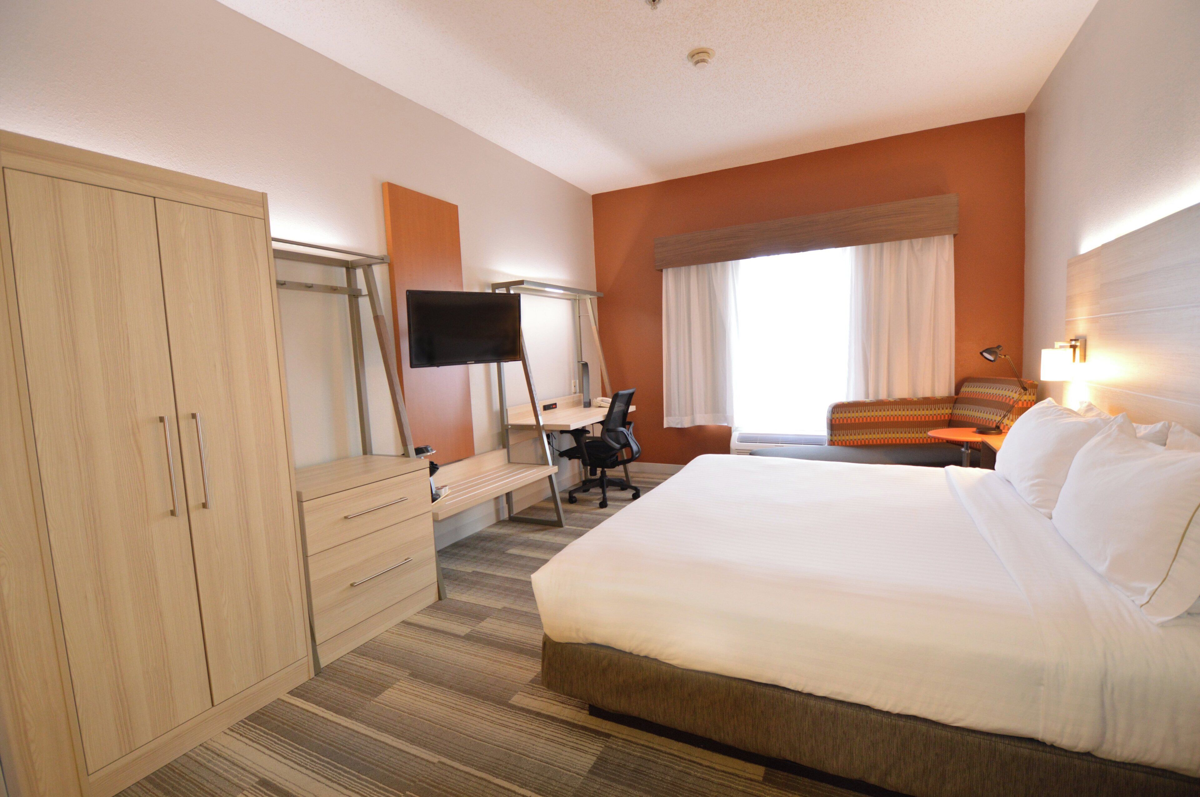 A hotel room with a bed, desk, chair, TV, wardrobe, and a window with curtains.