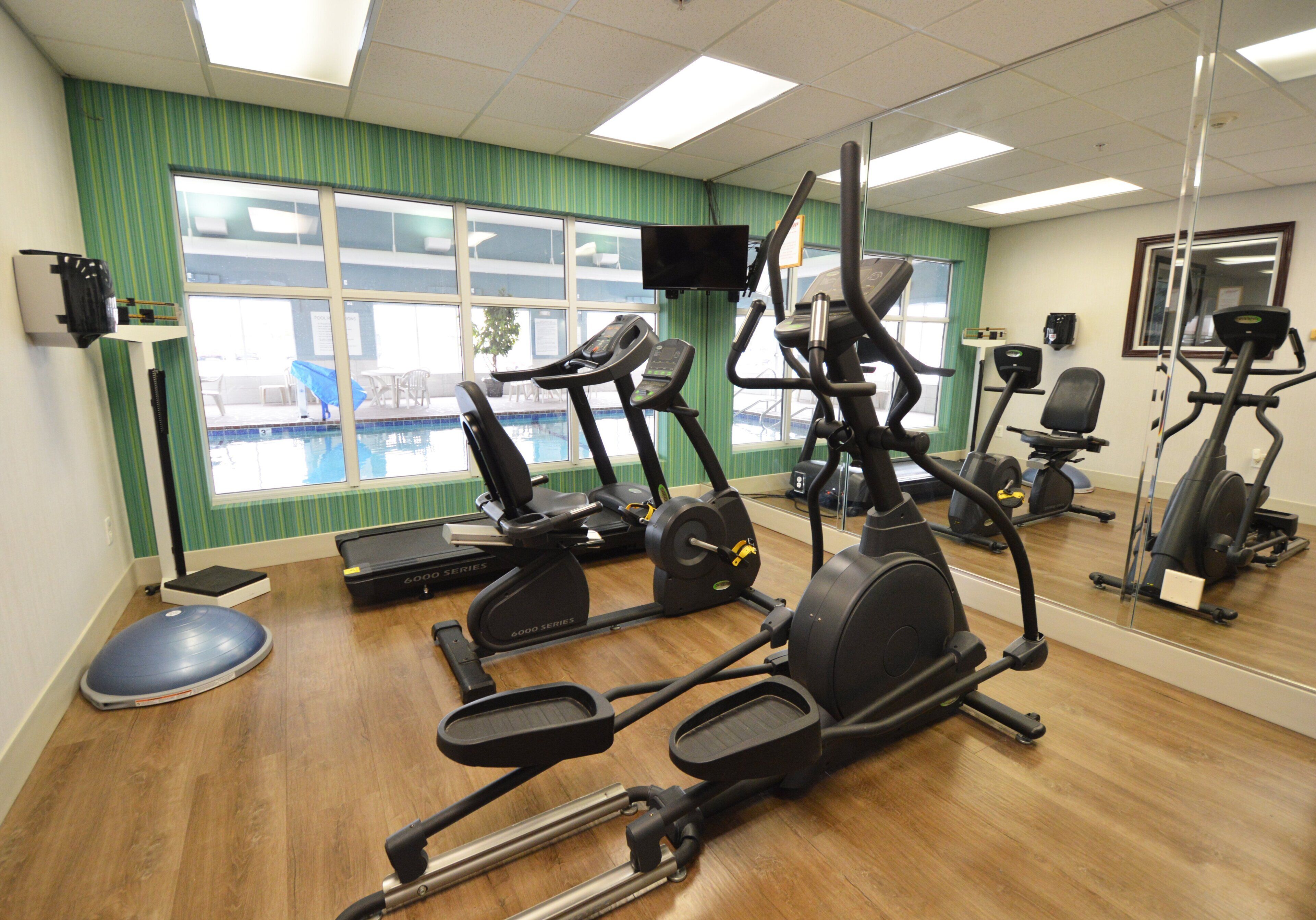 Un gimnasio con diversos equipos de ejercicio, una piscina visible a través de las ventanas y un espejo que refleja el equipo.