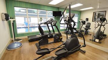 Un gimnasio con diversos equipos de ejercicio, una piscina visible a través de las ventanas y un espejo que refleja el equipo.