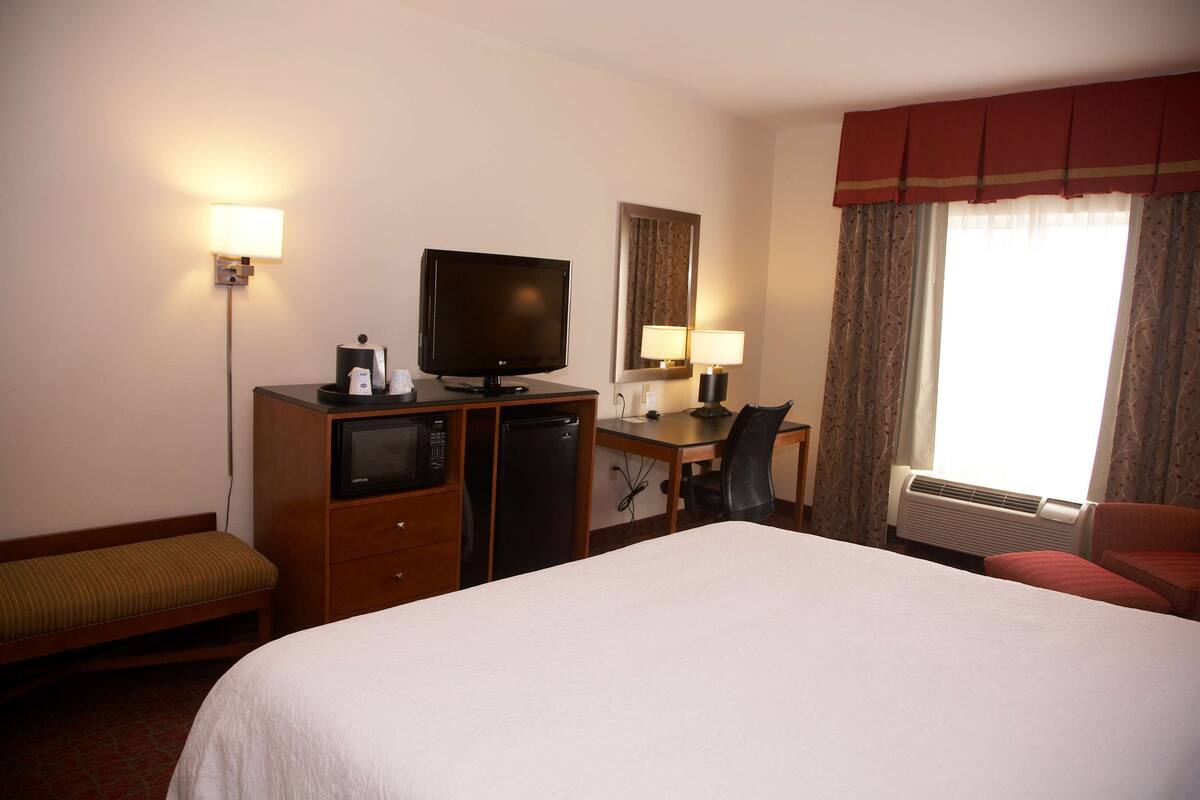 A hotel room with a bed, a desk with a computer, a television, a lamp, and a window with curtains.