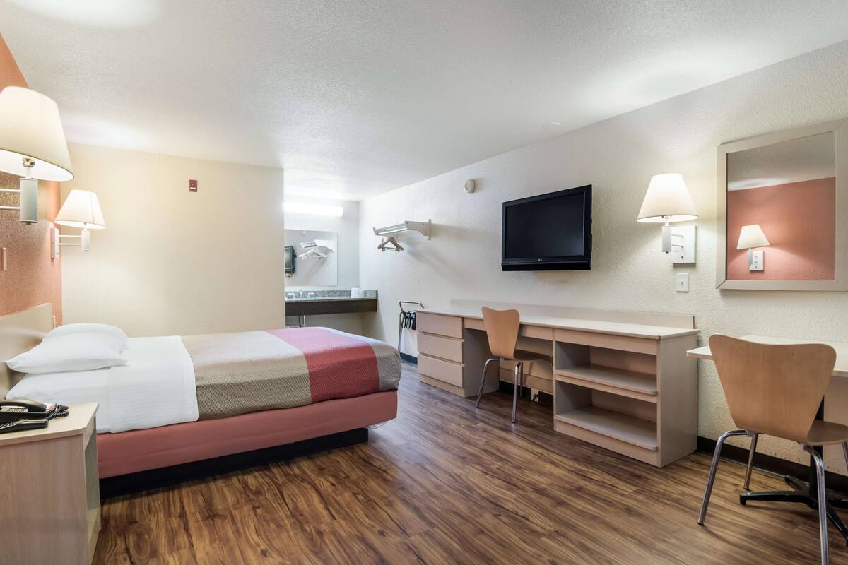 Habitación de hotel con cama, escritorio, silla, televisor y espejo.