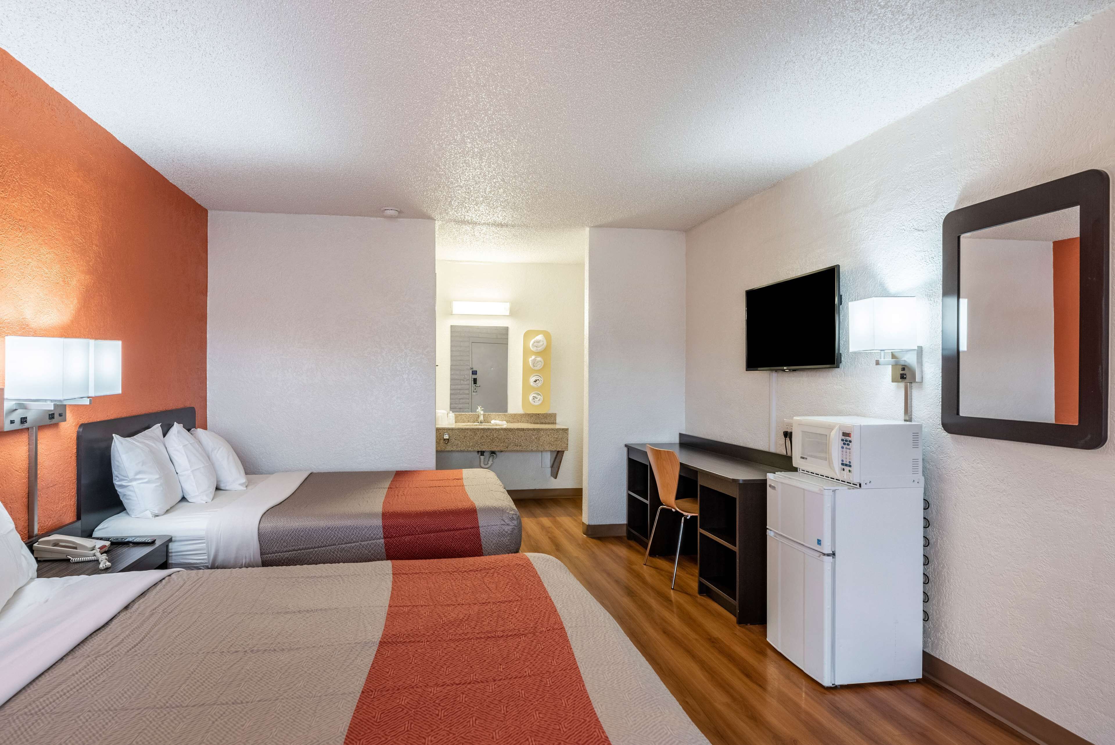 A hotel room with two beds, a desk, a refrigerator, a TV, and a bathroom visible in the background.