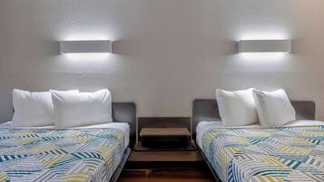 Habitación doble con dos camas, cada una con ropa blanca y almohadas, un pequeño banco entre las camas e iluminación en la pared.