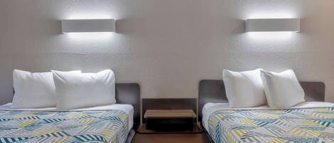 Habitación doble con dos camas, cada una con ropa blanca y almohadas, un pequeño banco entre las camas e iluminación en la pared.