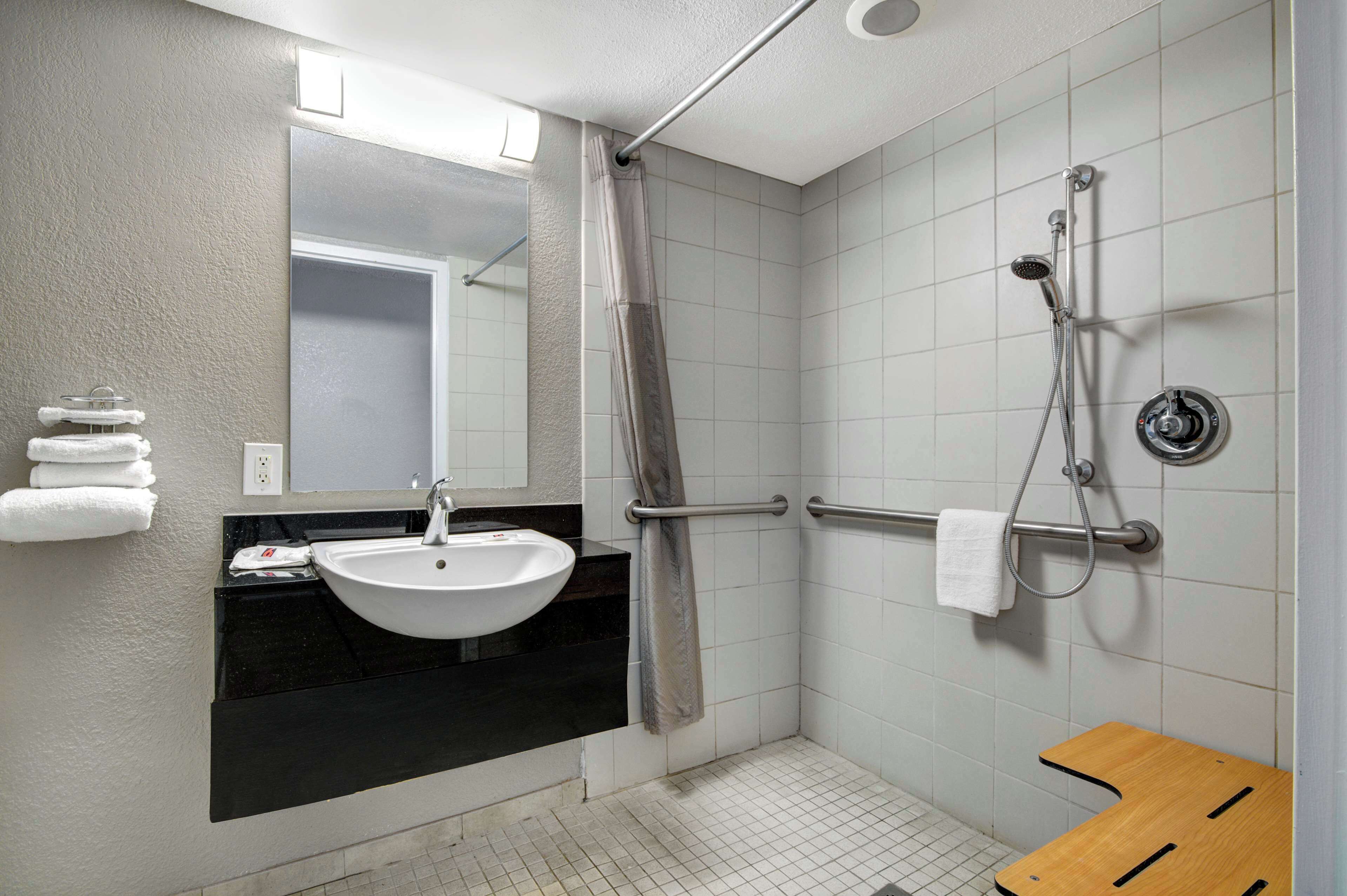 A modern bathroom with a white sink, a mirror, a shower with a handheld showerhead, and a wooden bench.