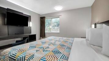 A hotel room with a bed, a flat-screen TV mounted on the wall, a window with curtains, and a desk with a chair.