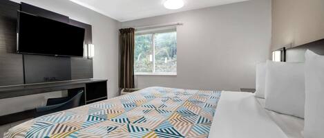 A hotel room with a bed, a flat-screen TV mounted on the wall, a window with curtains, and a desk with a chair.
