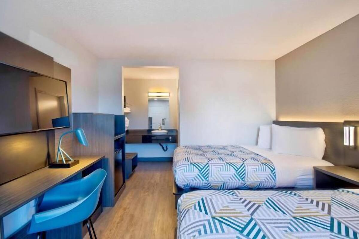 A hotel room with a desk, two beds with patterned bedding, a chair, and a mirror.