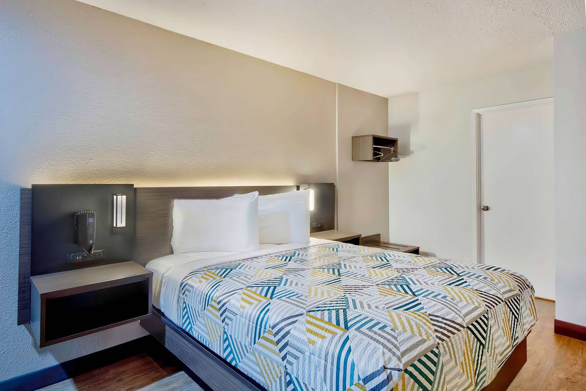 A modern hotel room with a neatly made bed, a bedside table with a lamp, and a small shelf above the bed.