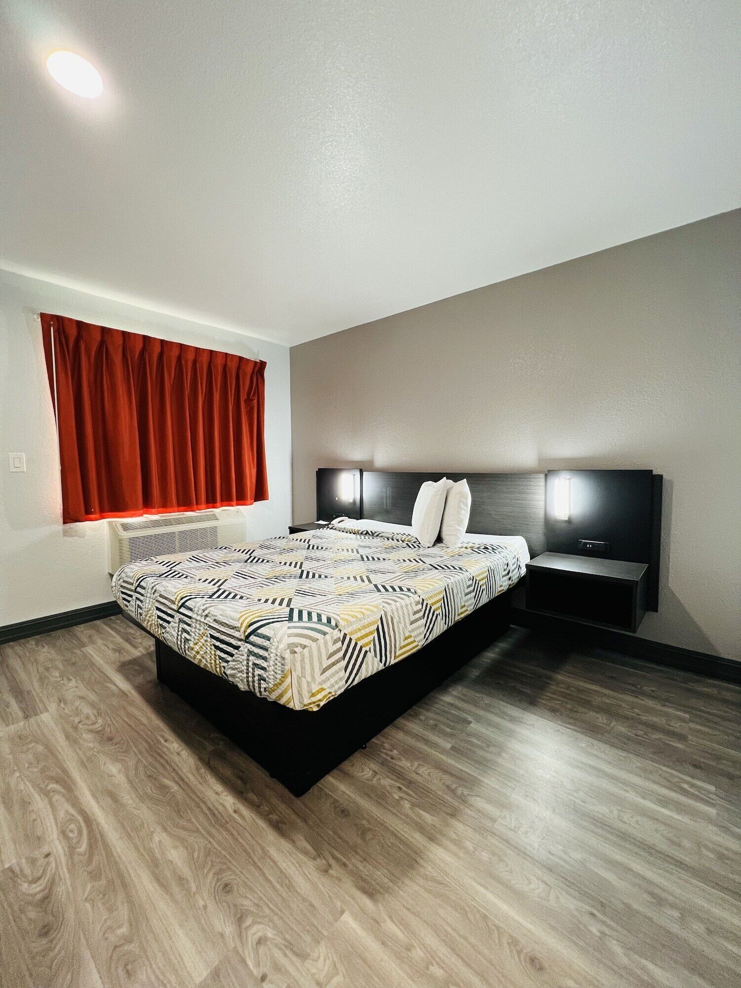 Un dormitorio moderno con una cama grande, una mesita de noche, un televisor en la pared y una ventana con cortinas rojas.
