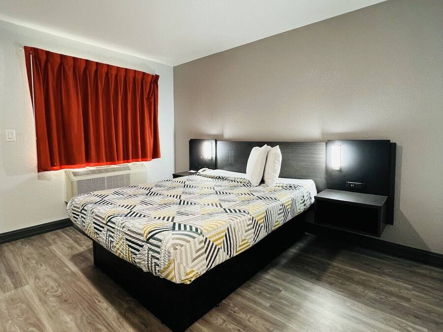 A modern bedroom with a large bed, a nightstand, a wall-mounted TV, and a window with red curtains.