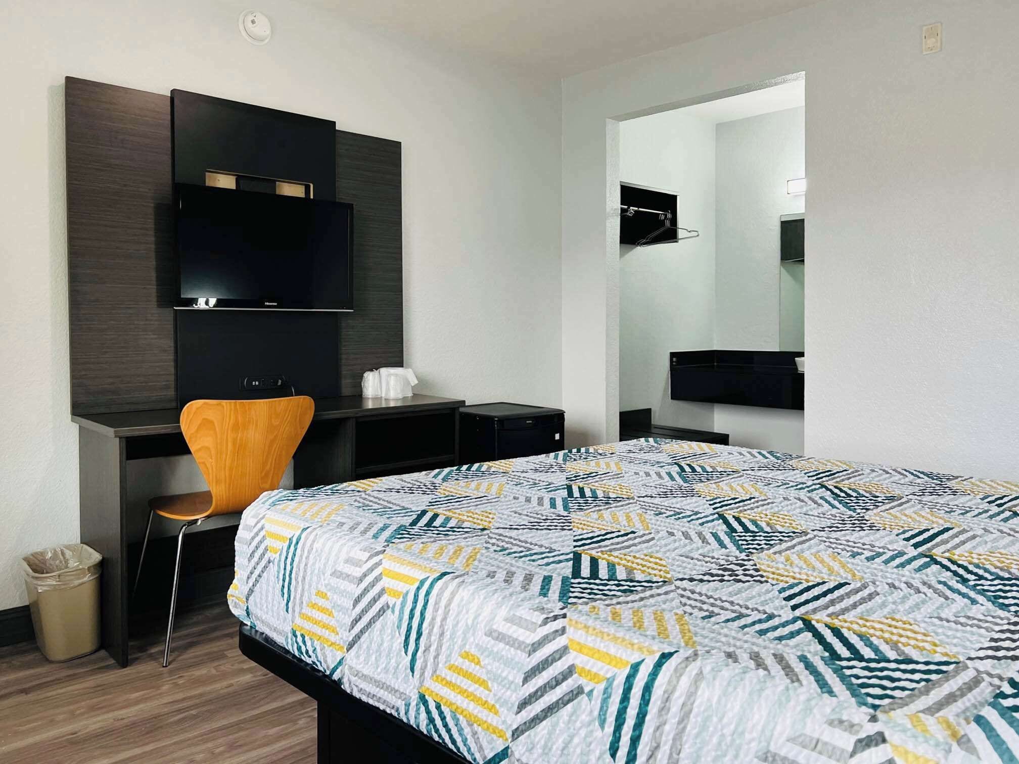 A hotel room with a bed, a desk with a chair, a television mounted on the wall, and a mirror above the desk.