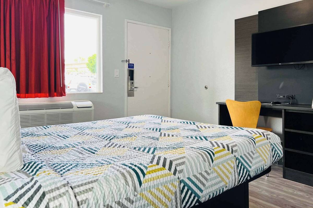 Una habitación de hotel con cama, escritorio con silla, televisor y ventana con cortinas.