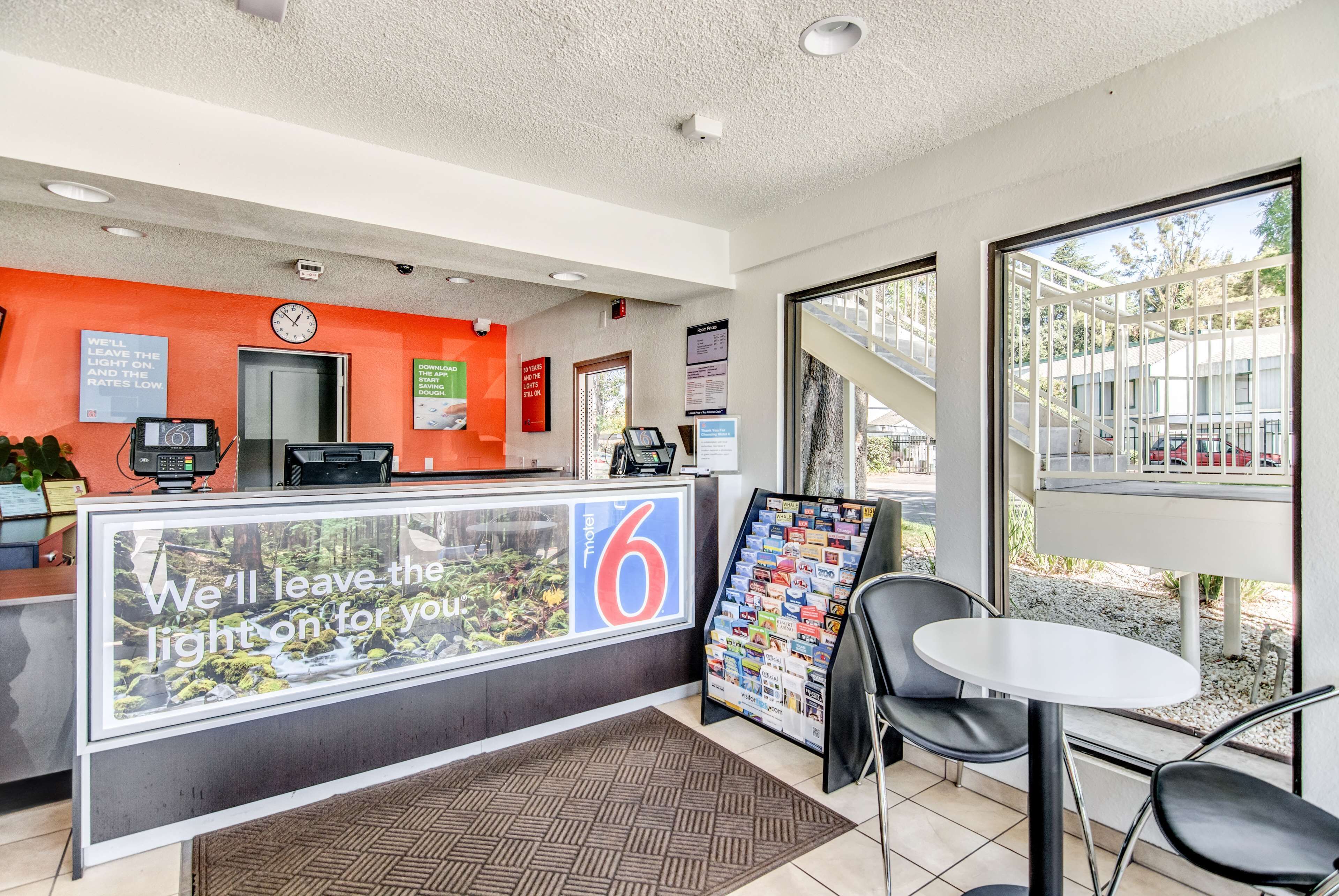 A hotel reception area with a large sign reading "We'll leave the light on for you," a reception desk, a clock, and a view of the outside through large windows.