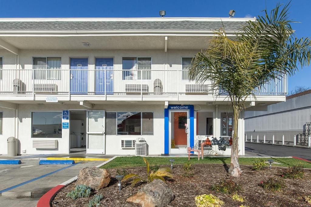A two-story motel with a 'welcome' sign, a covered parking area, and a small palm tree in the front yard.