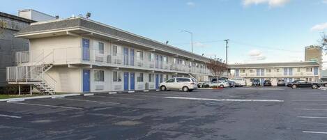A motel with a row of rooms, a parking lot, and a building in the background.