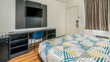 A hotel room with a bed, a desk with a chair, a television mounted on the wall, a door, and a window with curtains.